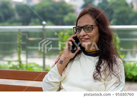 Mid Adult Smiling Woman with long brown hair Speaking on mobile phone outdoors. Closeup. She is wearing White T-shirt and specs. Front view. Head and Shoulder Shot. Garden lake side in the background. Mid Adult Smiling Woman with long brown hair Speaking on mobile phone outdoors. Closeup. She is wearing White T-shirt and specs. Front view. Head and Shoulder Shot. Garden lake side in the background. 95880760