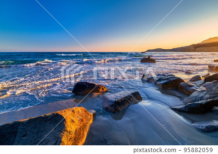Beautiful seascape of low tidal waves, clear sunset sky, hazy hills. Clear blue and azure waters approaching washed stones with little white foam. Beautiful seascape of low tidal waves, clear sunset sky, hazy hills. Clear blue and azure waters approaching washed stones with little white foam. 95882059