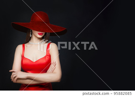 Young brunette woman with red lips makeup and diamond necklace and earrings and in burgundy wide hat, portrait on black background 95882085