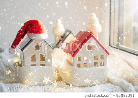 Cozy house is wrapped in a hat and scarf in a snowstorm -window sill decor. Winter, snow - home insulation, protection from cold and bad weather, room heating system. Festive mood, Christmas, New Year 95882391