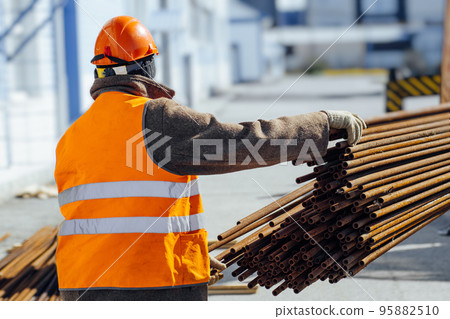 Slinger in construction helmet and orange vest unloads cargo on construction site. View from back 95882510