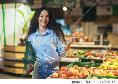 Portrait of happy and smiling hispanic female shopper in supermarket, woman smiling and looking at camera chooses apples and puts in eco bag. Portrait of happy and smiling hispanic female shopper in supermarket, woman smiling and looking at camera chooses apples and puts in eco bag. 95884047