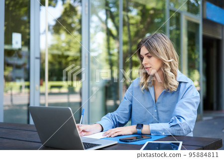 Successful businesswoman working outdoors, sitting on fresh air with laptop, documents and papers, looking at computer screen Successful businesswoman working outdoors, sitting on fresh air with laptop, documents and papers, looking at computer screen 95884811