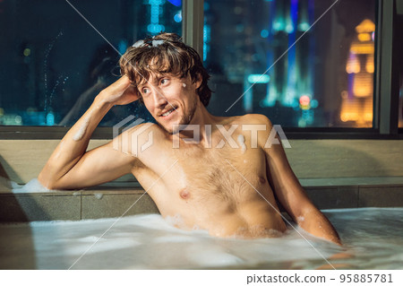 A young man takes a bath against a panoramic window with a view of the night city A young man takes a bath against a panoramic window with a view of the night city 95885781