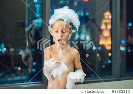 Happy little baby boy sitting in bath tub in the evening before going to sleep on the background of a window overlooking the evening city. Portrait of baby bathing in a bath full of foam near window 95885810