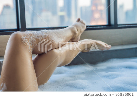 Young woman sits in a bath with foam and frangipani flowers against the background of a panoramic window overlooking the skyscrapers and a big evening city Young woman sits in a bath with foam and frangipani flowers against the background of a panoramic window overlooking the skyscrapers and a big evening city 95886001