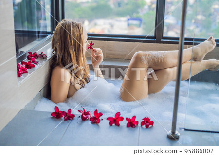 Young woman sits in a bath with foam and frangipani flowers against the background of a panoramic window overlooking the skyscrapers and a big evening city Young woman sits in a bath with foam and frangipani flowers against the background of a panoramic window overlooking the skyscrapers and a big evening city 95886002