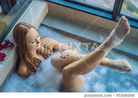 Young woman sits in a bath with foam and frangipani flowers against the background of a panoramic window overlooking the skyscrapers and a big evening city 95886006