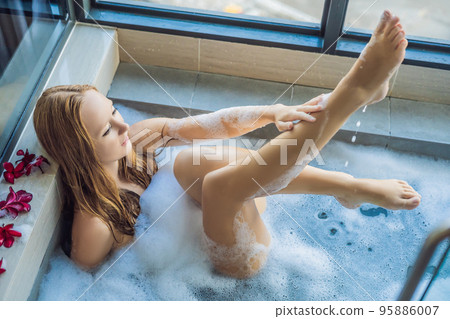 Young woman sits in a bath with foam and frangipani flowers against the background of a panoramic window overlooking the skyscrapers and a big evening city 95886007