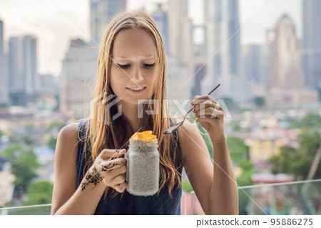 Young woman with mehendi from henna on hand eating chia pudding on her balcony overlooking the big city 95886275