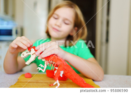 A blonde little girl makes a dinosaur craft out of wood and air plasticine.  95886590
