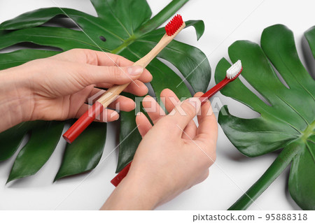Woman holding natural bamboo and plastic toothbrushes above tropical leaves on white background, closeup Woman holding natural bamboo and plastic toothbrushes above tropical leaves on white background, closeup 95888318