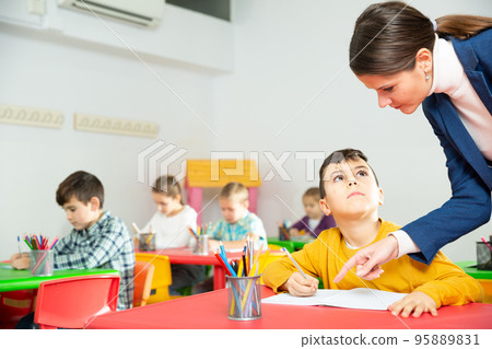 Friendly teacher woman helping boy during lesson in schoolroom Friendly teacher woman helping boy during lesson in schoolroom 95889831