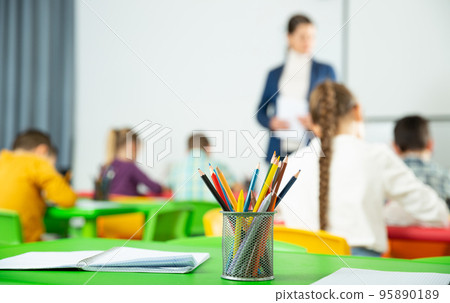 Stand with colored pencils and notebooks on the desk against the background of elementary school students Stand with colored pencils and notebooks on the desk against the background of elementary school students 95890189