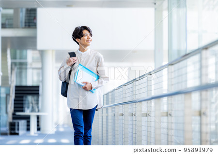Young male college student standing with notebook in campus corridor Young male college student standing with notebook in campus corridor 95891979