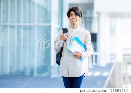 Young male college student standing with notebook in campus corridor Young male college student standing with notebook in campus corridor 95891984
