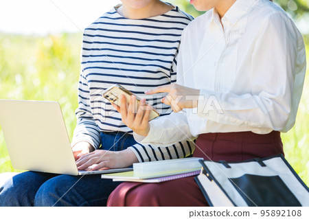 Two young female university students operating a smartphone and a personal computer in the park Two young female university students operating a smartphone and a personal computer in the park 95892108