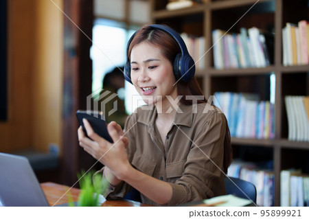 Portrait of a smiling Asian teenage girl wearing headphones and using a computer for online video conferencing in a library. Portrait of a smiling Asian teenage girl wearing headphones and using a computer for online video conferencing in a library. 95899921