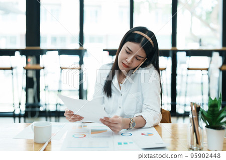 A portrait of a beautiful Asian female employee showing a stressed face while using the phone and financial documents on her desk. 95902448