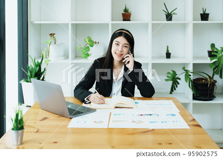 Portrait of a young Asian woman showing a smiling face as she uses her phone, computer and financial documents on her desk in the early morning hours. Portrait of a young Asian woman showing a smiling face as she uses her phone, computer and financial documents on her desk in the early morning hours. 95902575