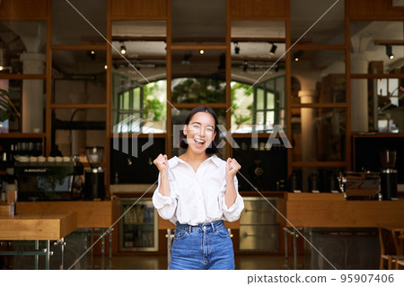 Happy enthusiastic asian girl, business owner triumphing, opening her own restaurant, standing in front of empty cafe 95907406