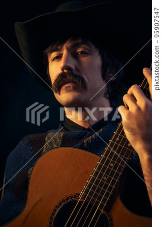 Portrait of brutal man with moustaches in big vintage scarf and black hat posing with guitar isolated over dark blue background. Deep look Portrait of brutal man with moustaches in big vintage scarf and black hat posing with guitar isolated over dark blue background. Deep look 95907547