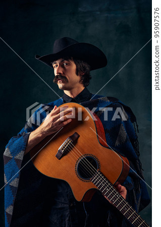 Portrait of man with moustaches in country style clothes playing guitar isolated over dark blue background. Affection and passion Portrait of man with moustaches in country style clothes playing guitar isolated over dark blue background. Affection and passion 95907576