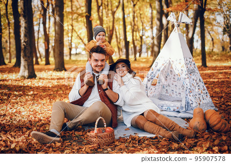 Family have fun in a autumn park. Cute blode in a white sweater. Parents with little son. Family sitting near wigwam; 95907578