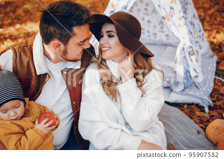 Family have fun in a autumn park. Cute blode in a white sweater. Parents with little son. Family sitting near wigwam; 95907582
