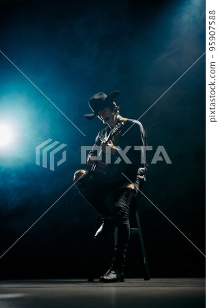 Portrait of man with moustaches in country style clothes playing guitar isolated over dark blue background with smoke. Live performance Portrait of man with moustaches in country style clothes playing guitar isolated over dark blue background with smoke. Live performance 95907588