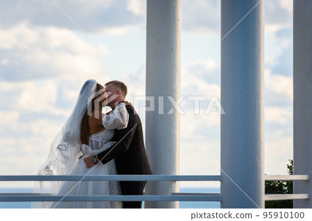 The newlyweds happily kiss in a beautiful picturesque gazebo against the sky 95910100