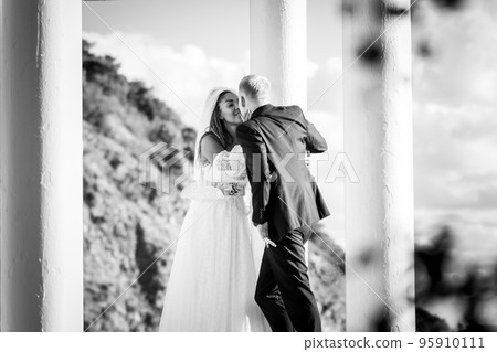 Happy newlyweds kiss in a beautiful gazebo with columns against the background of foliage and sky, black and white version 95910111