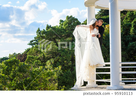 The newlyweds happily embrace in a beautiful gazebo with columns against the backdrop of foliage and sky The newlyweds happily embrace in a beautiful gazebo with columns against the backdrop of foliage and sky 95910113