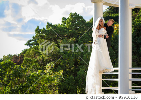 Happy newlyweds stand on the metal fence of the gazebo and look into the distance 95910116