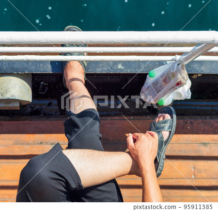 Cross legged man sitting on a sea side passage of a ferry boat with plastic bag hanged on handrail containing newspaper 95911385