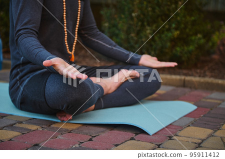 Cropped view of male athlete, yogi wearing rosary beads and gray sportswear, sitting on fitness mat in lotus position and palms up on knees, practicing yoga. Prayer, gratitude. Yoga pose. Meditation. 95911412