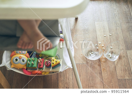 A small baby on a high chair dropped a cup of noodle soup on the wooden floor. The transparent plate is divided into two parts. Noodles in the form of figures. The table is dirty. High quality photo 95911564