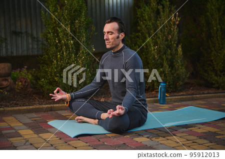 Handsome peaceful confident male athlete, a man yogi with rosary beads, meditating in lotus pose on a yoga mat at sunset. Sunset sunlight. Yoga practice. Meditation. Mindfulness. Enlightenment 95912013