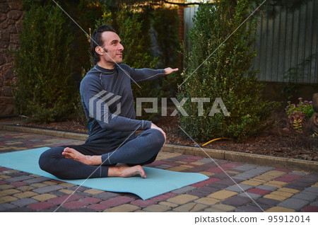Handsome active male athlete stretching body while sitting on fitness mat in lotus position, extending arms to sides during outdoor yoga practice at sunset. Health and body care concept. Sport. Yoga 95912014
