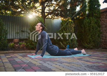 Active sporty man athlete, a sportsman exercising on a fitness mat, doing stretching exercises outdoors at sunset. Sunbeams fall on the backyard while an athletic guy working out on the house backyard Active sporty man athlete, a sportsman exercising on a fitness mat, doing stretching exercises outdoors at sunset. Sunbeams fall on the backyard while an athletic guy working out on the house backyard 95912017