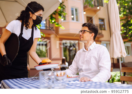 Waitress in protective mask serving delicious food for man in cozy restaurant. Waitress in protective mask serving delicious food for man in cozy restaurant. 95912775