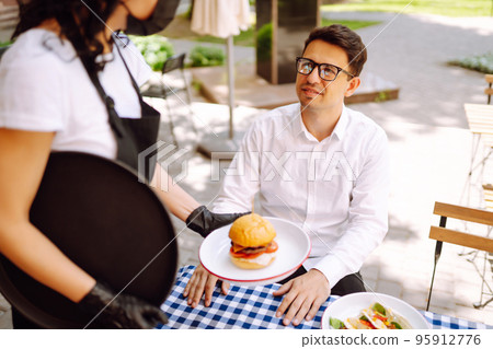 Waitress in protective mask serving delicious food for man in cozy restaurant. Waitress in protective mask serving delicious food for man in cozy restaurant. 95912776