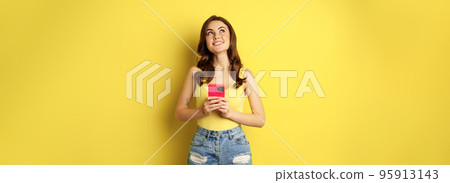 Technology and cellphone. Beautiful stylish girl order in online store, praying in application, using smartphone app and thinking, making decision, standing over yellow background Technology and cellphone. Beautiful stylish girl order in online store, praying in application, using smartphone app and thinking, making decision, standing over yellow background 95913143