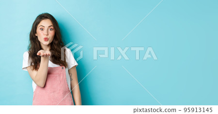 Beautiful girl blowing air kiss with romantic face, holding palm near lips and sending mwah at you, standing in summer outfit against blue background Beautiful girl blowing air kiss with romantic face, holding palm near lips and sending mwah at you, standing in summer outfit against blue background 95913145