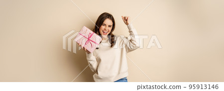 Happy smiling woman holding gift and triumphing, raising hand up joyfully, receive present on holiday, standing over beige background Happy smiling woman holding gift and triumphing, raising hand up joyfully, receive present on holiday, standing over beige background 95913146