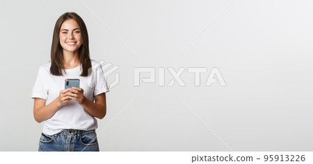 Attractive smiling brunette girl texting message, using mobile phone and looking at camera 95913226
