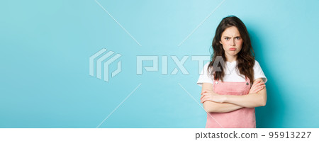 Portrait of angry and upset sulking girl, pouting about unfair situation, frowning and staring at camera, cross arms on chest defensive, standing over blue background 95913227