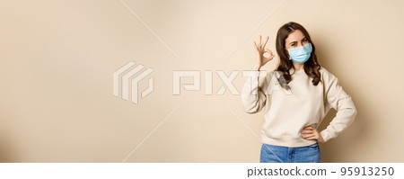 Covid-19, pandemic and quarantine concept. Young woman wears medical face mask during coronavirus omicron outbreak, showing okay sign, standing over beige background Covid-19, pandemic and quarantine concept. Young woman wears medical face mask during coronavirus omicron outbreak, showing okay sign, standing over beige background 95913250