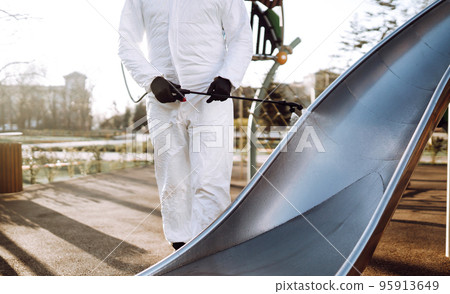Man wearing protective suit disinfecting the playground in the sun with spray. Covid -19. Cleaning. Man wearing protective suit disinfecting the playground in the sun with spray. Covid -19. Cleaning. 95913649