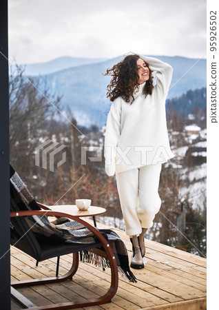 Curly young woman resting on terrace of modern barn house in the mountains. Happy female tourist enjoying in new cottage in winter. 95926502